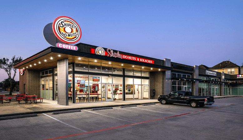 The exterior of a Shipley Do-Nuts store at dusk, with its glowing sign shining through large windows, hints at the sweet aroma within. Outside, a black pickup truck sits parked, emblematic of the brand's pipeline expansion that brings delightful treats to more communities.